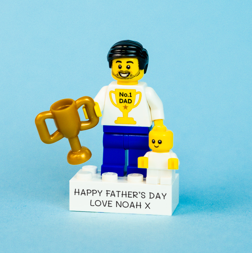 LEGO brick miniature figure holding a trophy and baby with 'Happy Father's Day' message on a blue background
