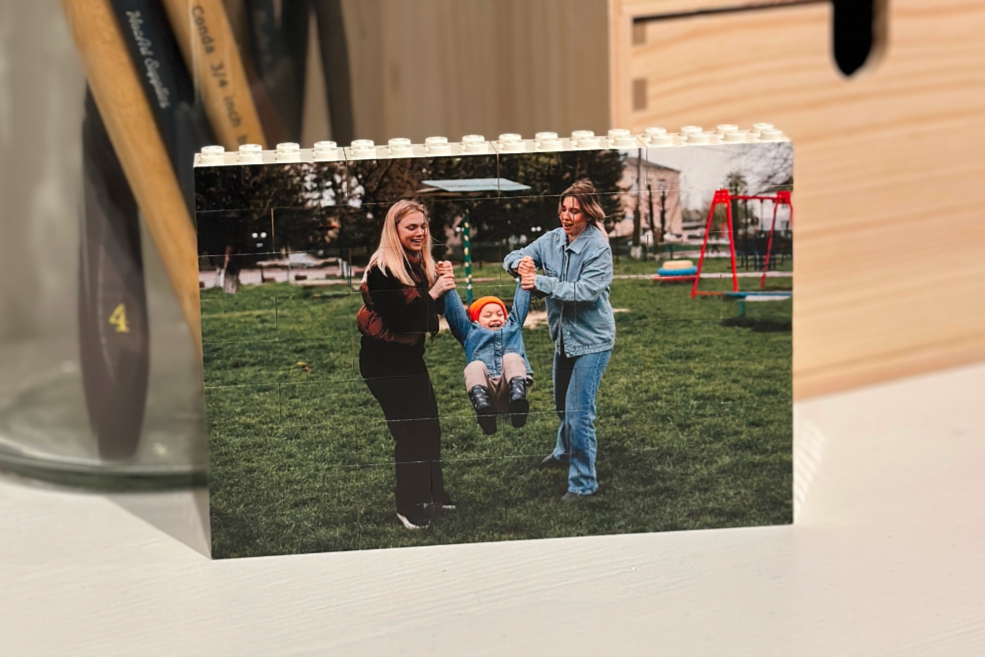 Small photo block made with LEGO bricks with 2 LQBTQ+ mums and their toddler on a surface with art paintbrushes on the desk in the background